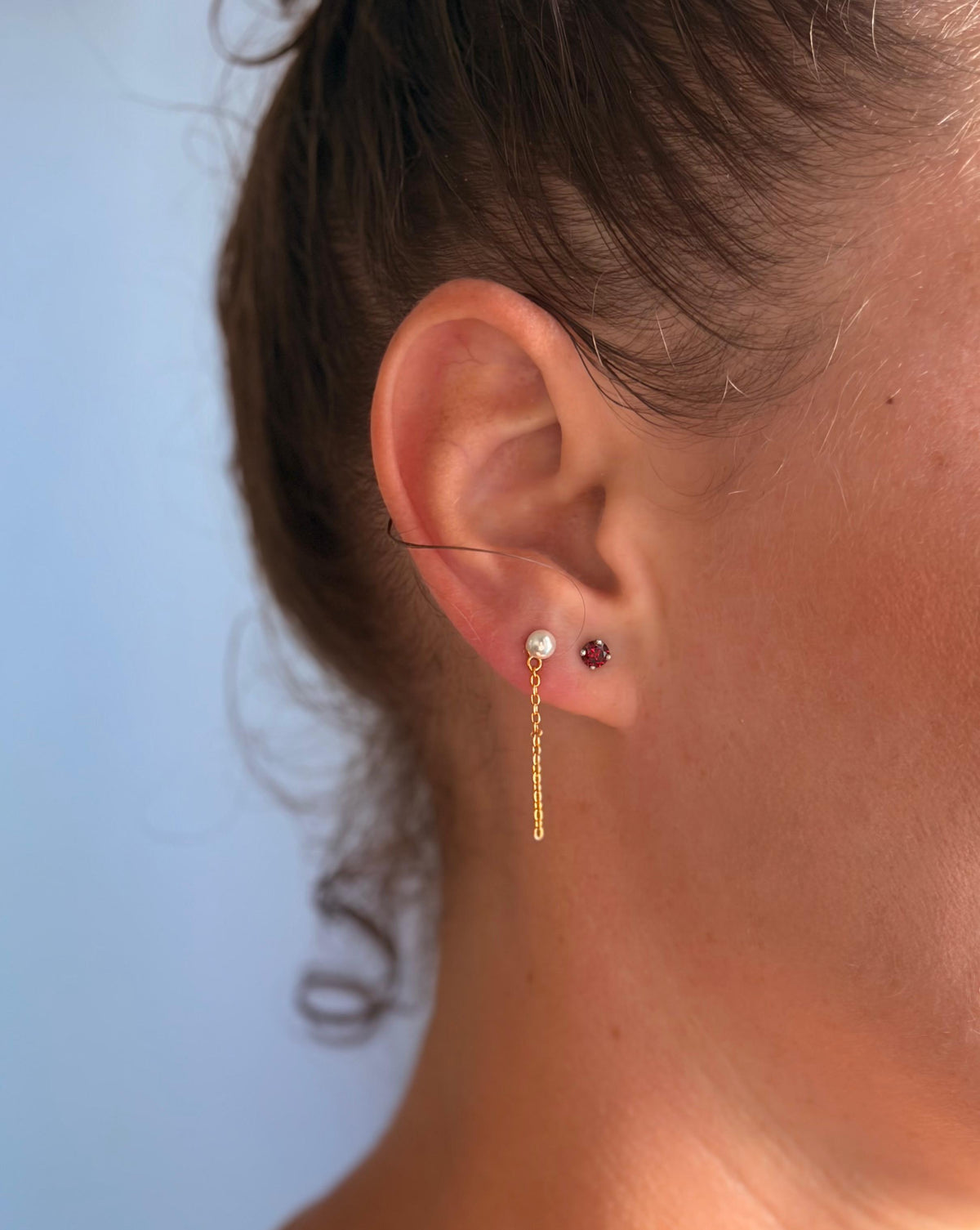 Femme portant les boucles d'oreilles sans fermoir Tina en Vermeil et Zéa en argent avec une pierre grenat, devant un fond uni bleuté